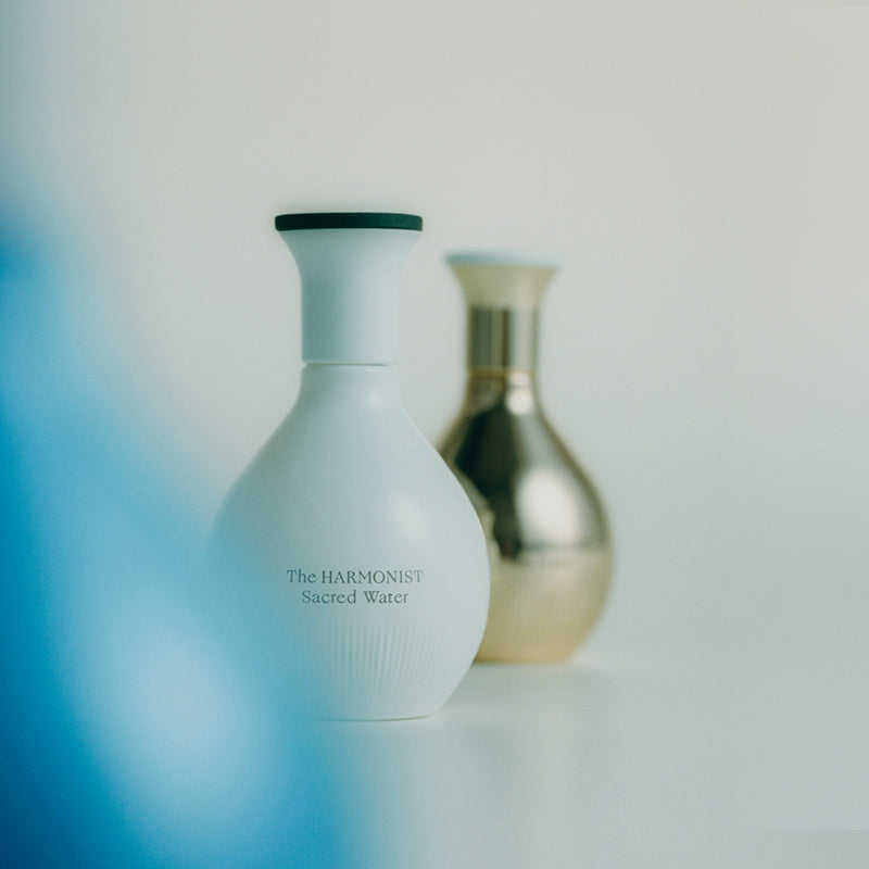 Minimalist photo of two elegant perfume bottles, one white labeled "The Harmonist Sacred Water" and one gold, set against a soft, light background.