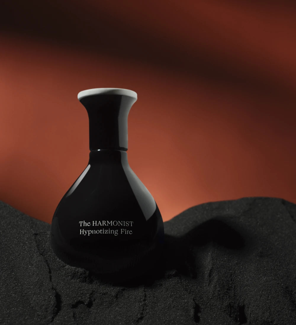 Sleek black bottle of “The Harmonist Hypnotizing Fire” perfume resting on black sand with a fiery red background.